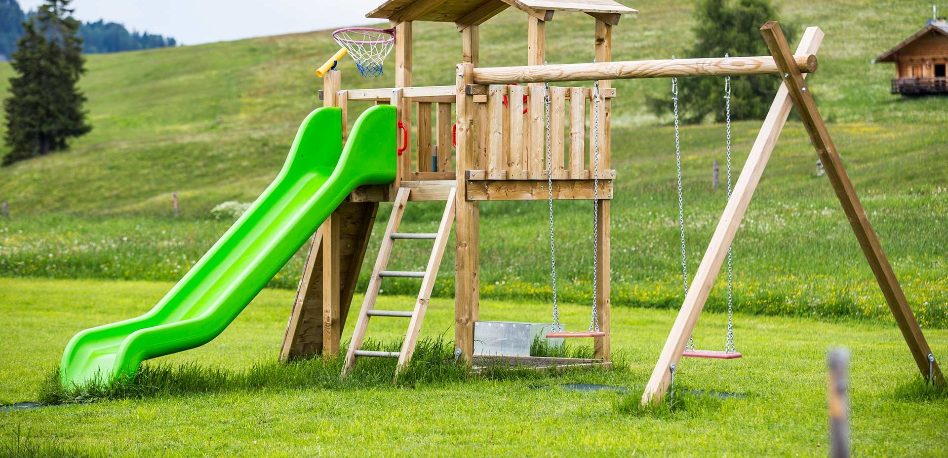 Ein Holzspielturm mit Rutsche und Schaukel auf einer großen weiten Wiese am Berg