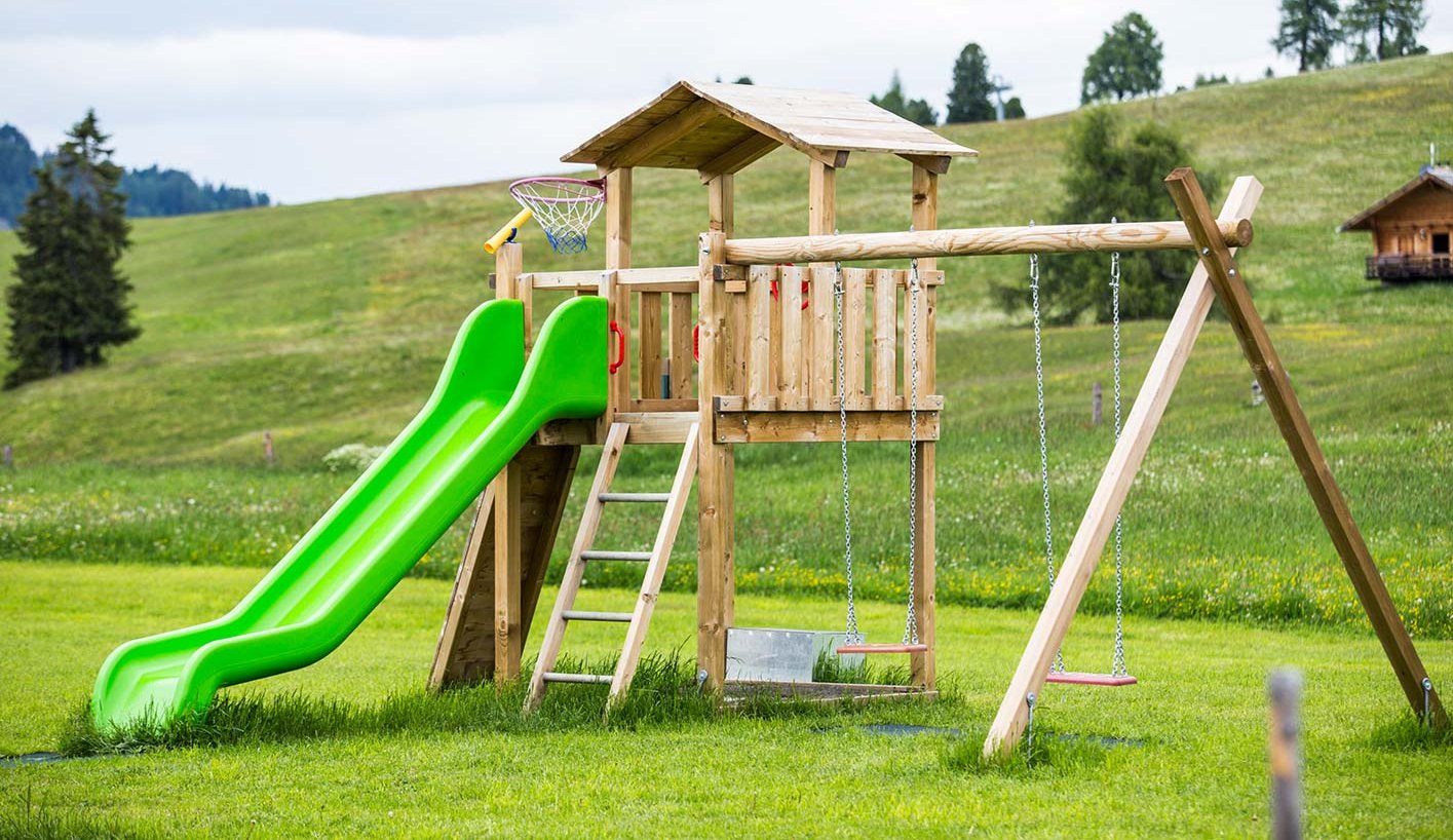 Ein Holzspielturm mit Rutsche und Schaukel auf einer großen weiten Wiese am Berg